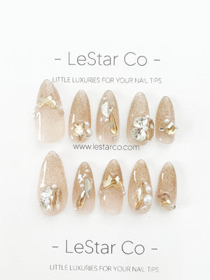Ten nude-colored, almond-shaped press-on nails with gold glitter and various embellishments, including rhinestones, pearls, and gold bows. The nails are arranged in two rows of five. The text "LeStar