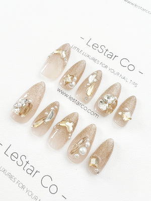 Set of ten nude-colored press-on nails with gold glitter and rhinestones. The nails are decorated with pearls and gold accents. The text "LeStar Co - Little Luxuries for Your Nail Tips" is printed on