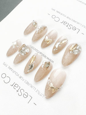 Set of ten nude-colored false nails with glitter and gold accents. Each nail is decorated with a different design featuring pearls, stars, and gold accents. The nails are arranged in a row on a white