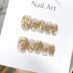 Set of ten beige press-on nails with a floral design featuring small yellow flowers and brown leaves. The nails are arranged in two rows on a white surface. "Nail Art." is written on the surface.