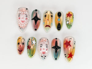 Ten false fingernails with different designs. Each nail is painted with a different design, including a face with a pearl necklace, a black star, a crying face, a black circle with a turquoise gem, a