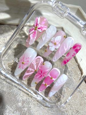 A set of ten pink and white artificial nails with floral designs. The nails are arranged in a clear plastic case. Some of the nails have gold accents.