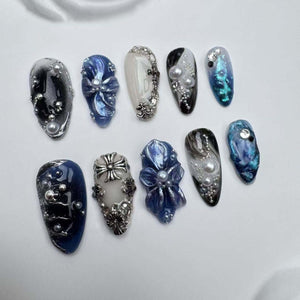 Ten blue and white nail tips with various designs, including a cross, a flower, and a swirl pattern. Each nail tip is decorated with pearls and rhinestones.