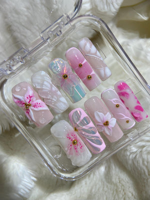 A set of ten pink and white artificial nails with floral designs and gold accents. The nails are arranged in a clear plastic case.