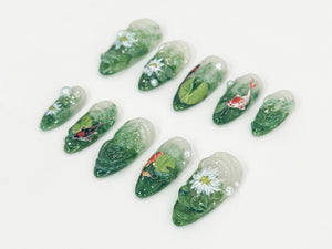 Ten clear nail tips with green and white designs. The designs include flowers, leaves, and fish.