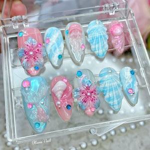 A set of ten false nails with a beach theme. The nails are decorated with seashells, flowers, and iridescent glitter. The nails are displayed in a clear plastic box.