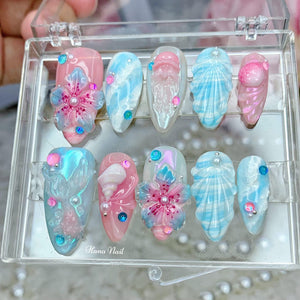 A set of ten false nails with various designs inspired by the ocean. The designs include seashells, flowers, and swirls. The nails are in a clear plastic case with the text "Hana Nail" printed on the