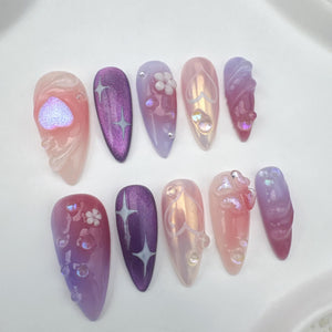 Set of ten false nails with a gradient design. The nails are in shades of pink, purple, and white. Some nails have a heart design, while others have a flower design.