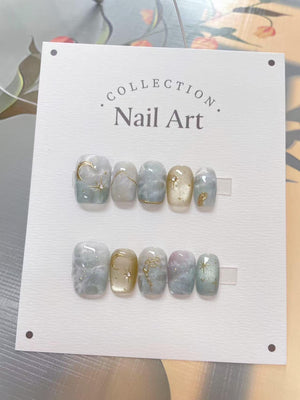 A set of press-on nails with a light blue and gold color scheme. The nails feature a variety of designs, including moons, stars, and abstract patterns. The text "COLLECTION Nail Art" is printed on the