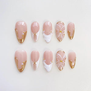 A set of ten almond-shaped false nails. The nails feature a pale pink base with gold accents, white designs, and floral embellishments. Some nails have a gold border, while others have a starburst or a flower design.