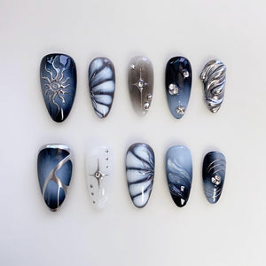 A set of ten artificial nails in various designs. The nails feature a range of colors, including black, white, and gray, with decorative elements such as silver swirls, starbursts, and rhinestones. The nails are oval-shaped.