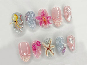 A set of ten artificial nails with various ocean-themed designs. The nails feature detailed artwork of an octopus, starfish, and flowers in shades of pink, white, and blue. Each nail has a glossy fini
