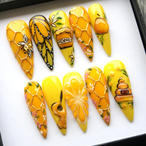 A set of ten yellow artificial nails with various designs. Some nails feature a honeycomb pattern, a butterfly, a bee, and a honey pot with the word "HONEY". Other nails have floral and fruit designs.