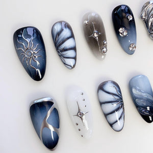 A collection of artificial nails with various designs. Some nails feature silver sunburst and floral patterns, while others have star and gem embellishments. The color palette includes shades of blue, gray, and white, creating a stylish and artistic