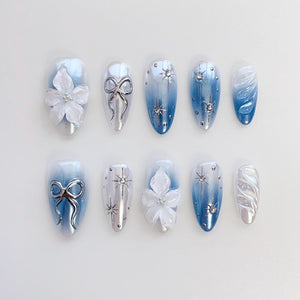 A set of ten artificial nails in various designs. Some nails feature a gradient of blue and white, while others have silver bows, starbursts, or white floral embellishments. The nails are oval-shaped and have a glossy finish.