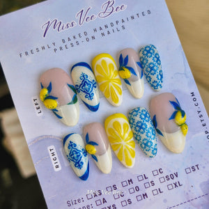 A set of hand-painted press-on nails with various designs. The nails feature a white base with yellow lemon slices, blue and white patterns, and lemon and leaf accents. The text "Miss Vee Bee" is visi