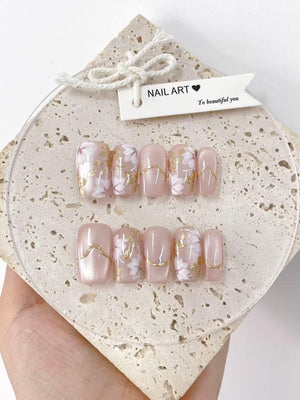 A set of artificial nails with a light pink base, adorned with white floral designs and gold accents. The nails are displayed on a textured, light-colored surface, with a tag that reads "NAIL ART" and