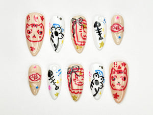 A set of ten almond-shaped artificial nails, each hand-painted with unique designs. The designs include a cat face, a koala, a fish skeleton, and abstract patterns in red, black, and blue on a neutral base.