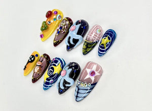A collection of ten almond-shaped, painted fingernails. Each nail features a unique design with various colors, including yellow, brown, blue, and pink. Embellishments include rhinestones and intricate patterns, showcasing nail art.