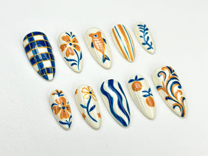 A collection of ten almond-shaped false nails, each hand-painted with unique designs. The designs include floral patterns, fish, stripes, and abstract swirls in shades of blue, orange, and white.