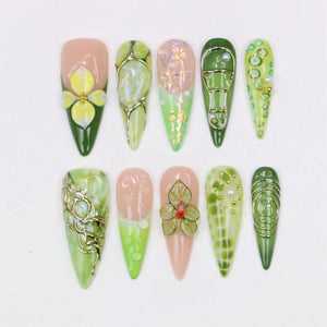 A collection of ten almond-shaped artificial nails with various designs. The nails feature a range of colors, including pink, green, and gold, with floral and abstract patterns. Some nails have 3D embellishments.