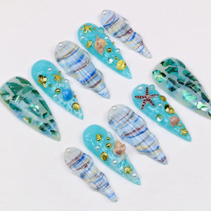 A collection of artificial nails with a beach theme. The nails are various shades of blue, decorated with seashells, starfish, gold accents, and faux pearls. Some nails have a marbled effect.