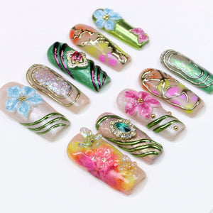 A collection of ten artificial nails, each showcasing a unique design. The nails feature floral and abstract patterns, with embellishments like glitter, gems, and metallic accents. Colors include pink, blue, green, and gold.
