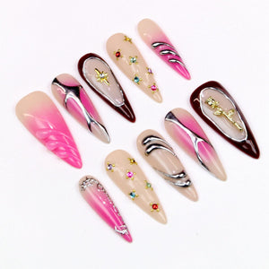 A collection of ten long, almond-shaped artificial nails. The nails feature various designs, including metallic accents, rhinestones, and floral embellishments. Colors include pink, nude, and burgundy.