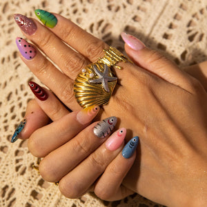 A hand with long, colorful, painted fingernails. The nails feature various designs, including fish, polka dots, and glitter. A gold ring with a seashell and starfish design is also visible.