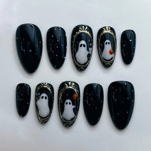 A set of ten almond-shaped press-on nails with a glossy black base featuring white speckles. Several nails display hand-painted white ghosts framed in metallic gold, with one ghost holding an orange b