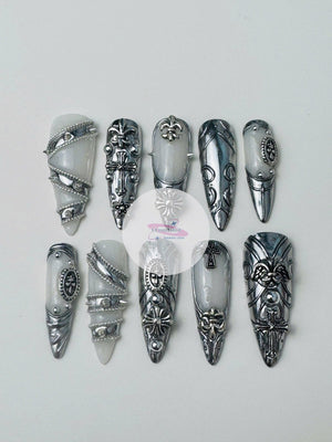 A set of ten long, silver and white press-on nails. Each nail features intricate silver embellishments, including crosses, fleur-de-lis, and ornate designs. The nails have a glossy finish and a pointe