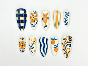 A set of ten almond-shaped press-on nails with various hand-painted designs. The designs include plaid, flowers, fish, stripes, and swirls in shades of blue, orange, and white. The nails are arranged