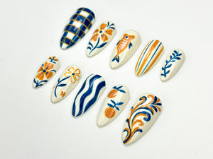A collection of ten press-on nails with various designs. The nails feature patterns such as plaid, flowers, fish, stripes, and swirls, in colors of blue, orange, and white. The nails are oval-shaped a