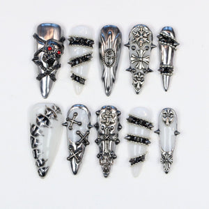 A collection of ten silver-colored, gothic-style nail art designs. Each nail features unique embellishments, including skulls, crosses, spikes, and geometric patterns. The nails are displayed against a white background.