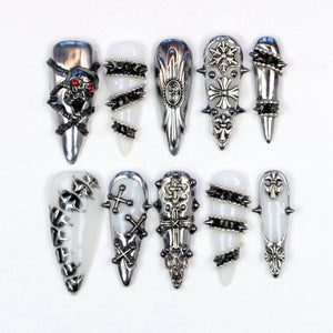 A collection of ten long, pointed nail designs. The nails feature silver and black embellishments, including skulls, crosses, spikes, and geometric patterns. Some nails have a white base, while others are silver.