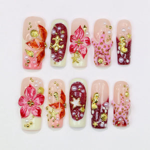 A set of ten artificial nails with various designs. Each nail features a base color of either nude or white, adorned with 3D embellishments like flowers, starfish, pearls, and gold accents. The designs incorporate shades of red, pink, and gold.