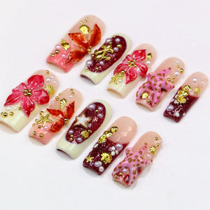 A collection of ten artificial nails, each showcasing a unique design. The nails feature a variety of embellishments, including gold-colored accents, pearls, and floral or seashell motifs. The color palette includes shades of pink, red, and cream.