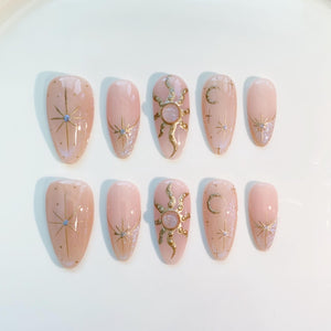 A set of ten almond-shaped press-on nails in a light pink hue. Each nail features gold celestial designs, including stars, suns, and moons, with glitter accents. The nails are arranged on a white surface.