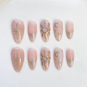 Set of ten almond-shaped false nails in a light pink hue. Each nail features celestial designs in gold and silver, including stars, suns, and moons. Some nails have a glittery accent at the base.