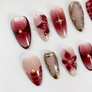 A collection of artificial nails with various designs. Some nails feature a gradient of red and pink hues, while others showcase 3D floral embellishments, gold star accents, and metallic gold frames. The nails are oval-shaped.