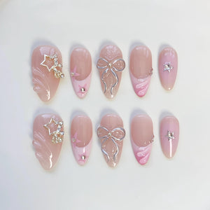 A set of ten artificial nails in various designs. The nails are primarily pink with decorative elements including silver bows, star-shaped rhinestones, and pink swirls. Some nails feature a gradient effect.