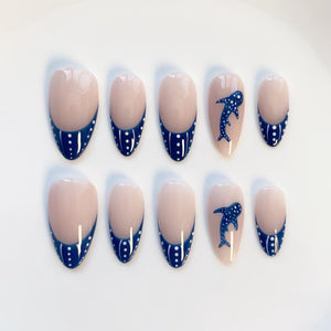 A set of ten oval-shaped false nails. The nails have a nude base with a navy blue French tip adorned with white polka dots. Two nails feature a navy blue whale design.