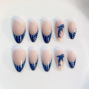 Set of ten artificial nails. Each nail has a nude base with a navy blue tip adorned with white polka dots. Two nails feature a navy blue whale design. The nails are oval-shaped.