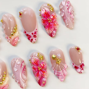 A collection of artificial nails with various designs. Some nails feature pink and white swirls, while others showcase 3D floral and seashell embellishments. Gold beads and gems add accents to the nails, creating a glamorous look.