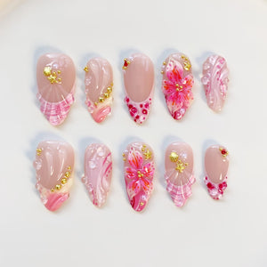 A set of ten almond-shaped artificial nails. Each nail features a nude base with pink and gold accents. Designs include floral patterns, glitter, and textured details. The nails are arranged on a white surface.
