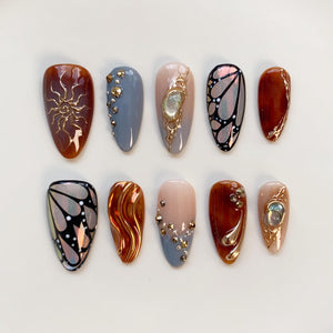 A collection of ten almond-shaped artificial nails. The nails feature various designs, including gold sunbursts, butterfly wings, and embellishments with gold and silver rhinestones. Colors include brown, gray, nude, and iridescent finishes.