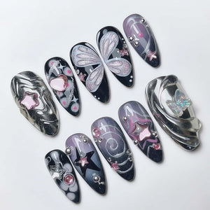 A collection of ten artificial nails with various designs. The nails feature a mix of colors including black, silver, pink, and purple. Some nails have star, heart, and butterfly designs, while others have swirls and embellishments.