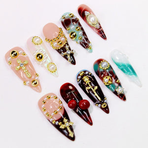 A collection of long, pointed artificial nails in various designs. Some nails feature gold crosses, pearls, and gold beads. Others have cherry accents, and a gradient of colors including red, pink, and teal.
