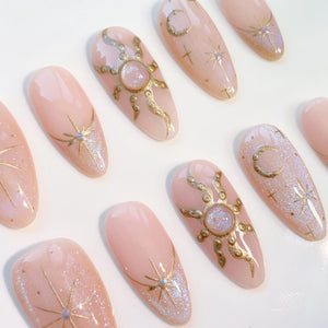 A collection of light pink, almond-shaped press-on nails. Some nails feature gold sun and moon designs, while others have gold starburst accents with glitter. The nails are arranged on a white surface.