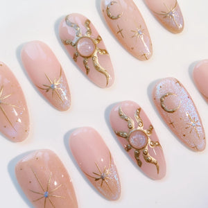 A collection of almond-shaped artificial nails with a light pink base. Each nail features celestial designs in gold, including suns, moons, and stars. Some nails have glitter accents, creating a shimmering effect. The overall aesthetic is elegant and